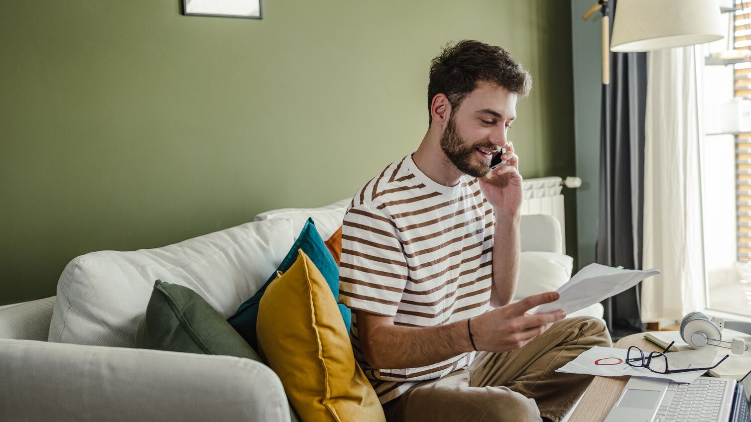 Young man sitting on the sofa and talking on the phone while holding documents