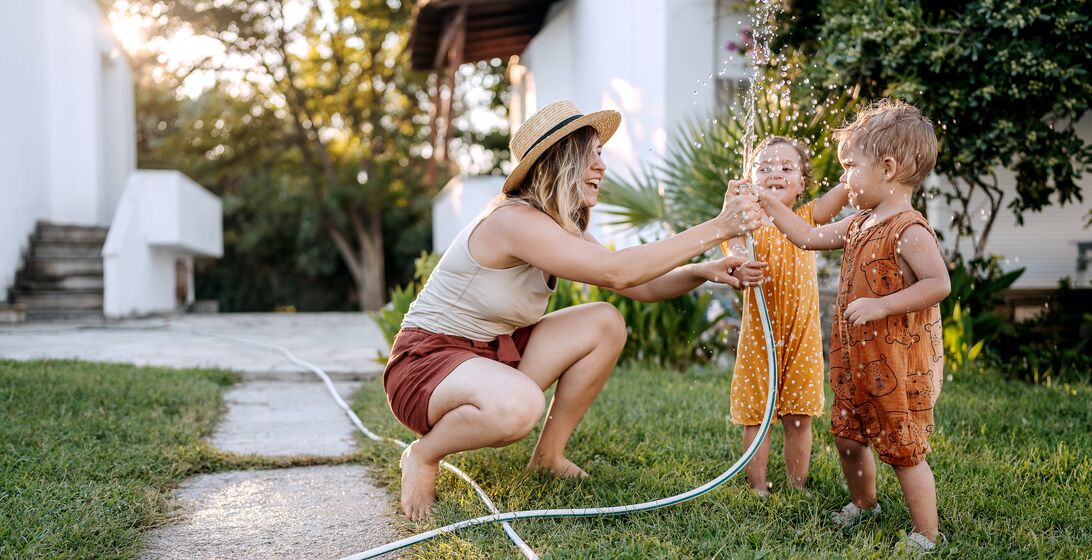 Mother and her cute little children playing with water with garden hose in a backyard