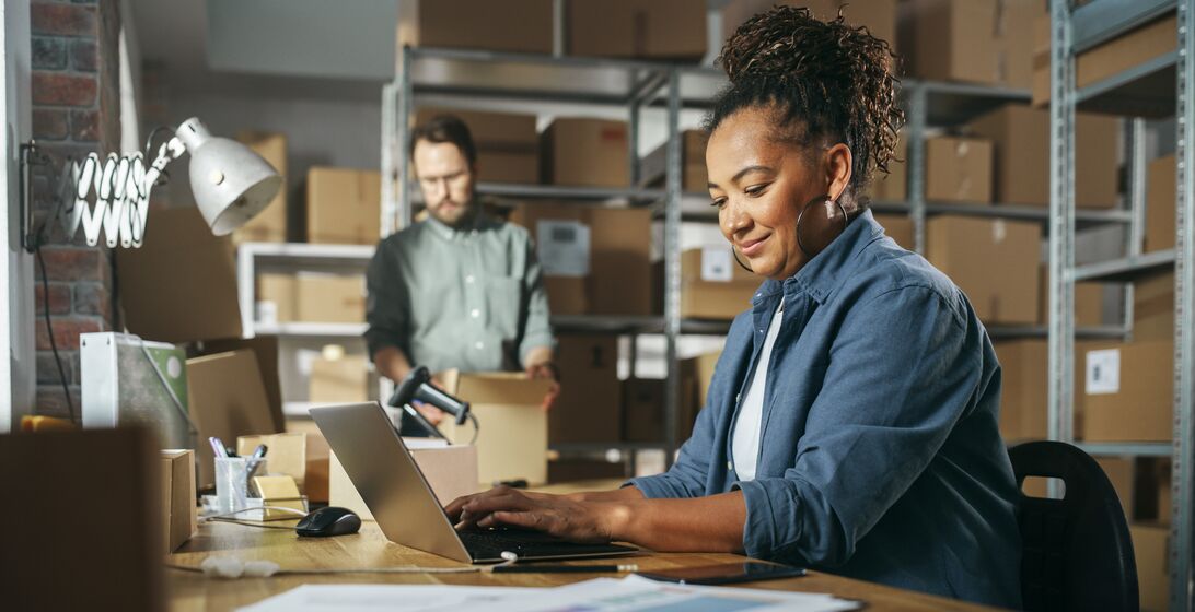 Diverse Male and Female Warehouse Inventory Managers Talking, Using Laptop Computer and Checking Retail Stock. Rows of Shelves Full of Cardboard Box Packages in the Background.