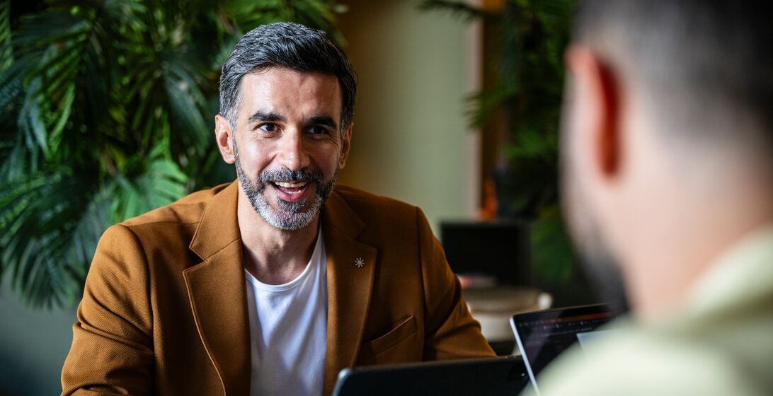 Man wearing a tan blazer sitting at a table across from another man wearing a sage green button up top