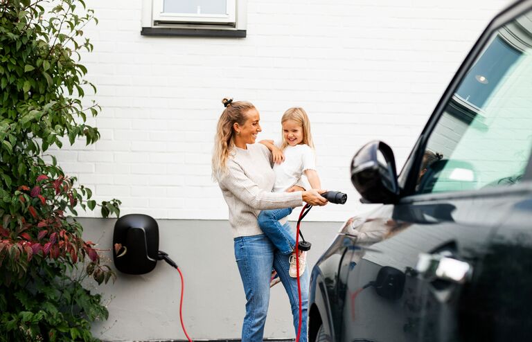 Happy woman carrying girl while walking towards electric car with charger