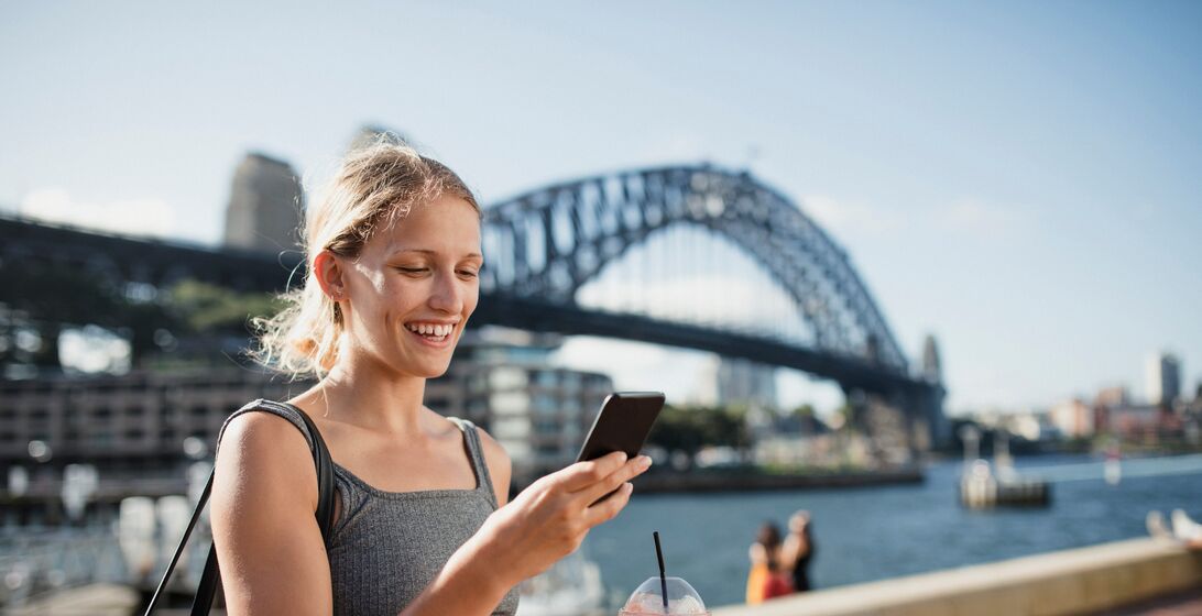 A woman uses her phone with a view of Sydney Harbour Bridge in the background.