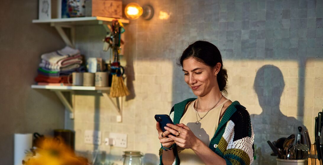 Smiling woman in her 30's standing in kitchen texting on smart phone, communication, wireless technology, the internet