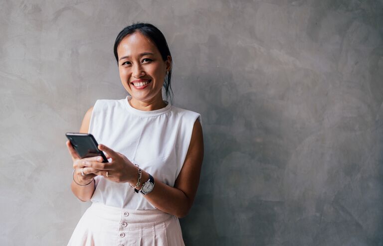 Portrait of a young Asian woman texting on her smartphone, looking at the camera, smiling.