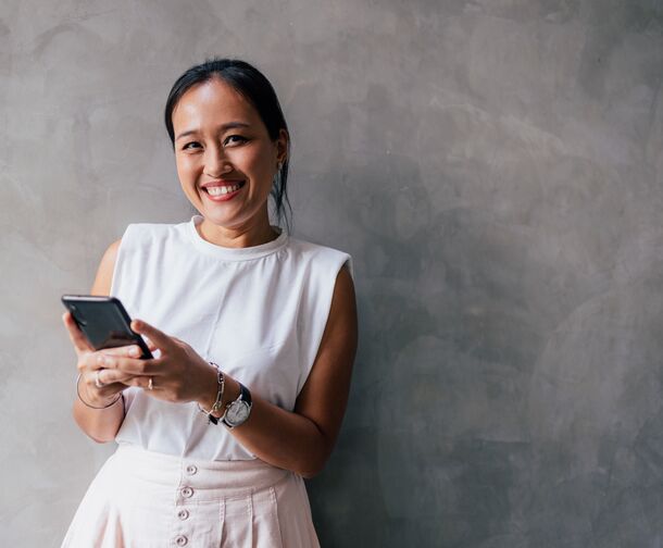 Portrait of a young Asian woman texting on her smartphone, looking at the camera, smiling.