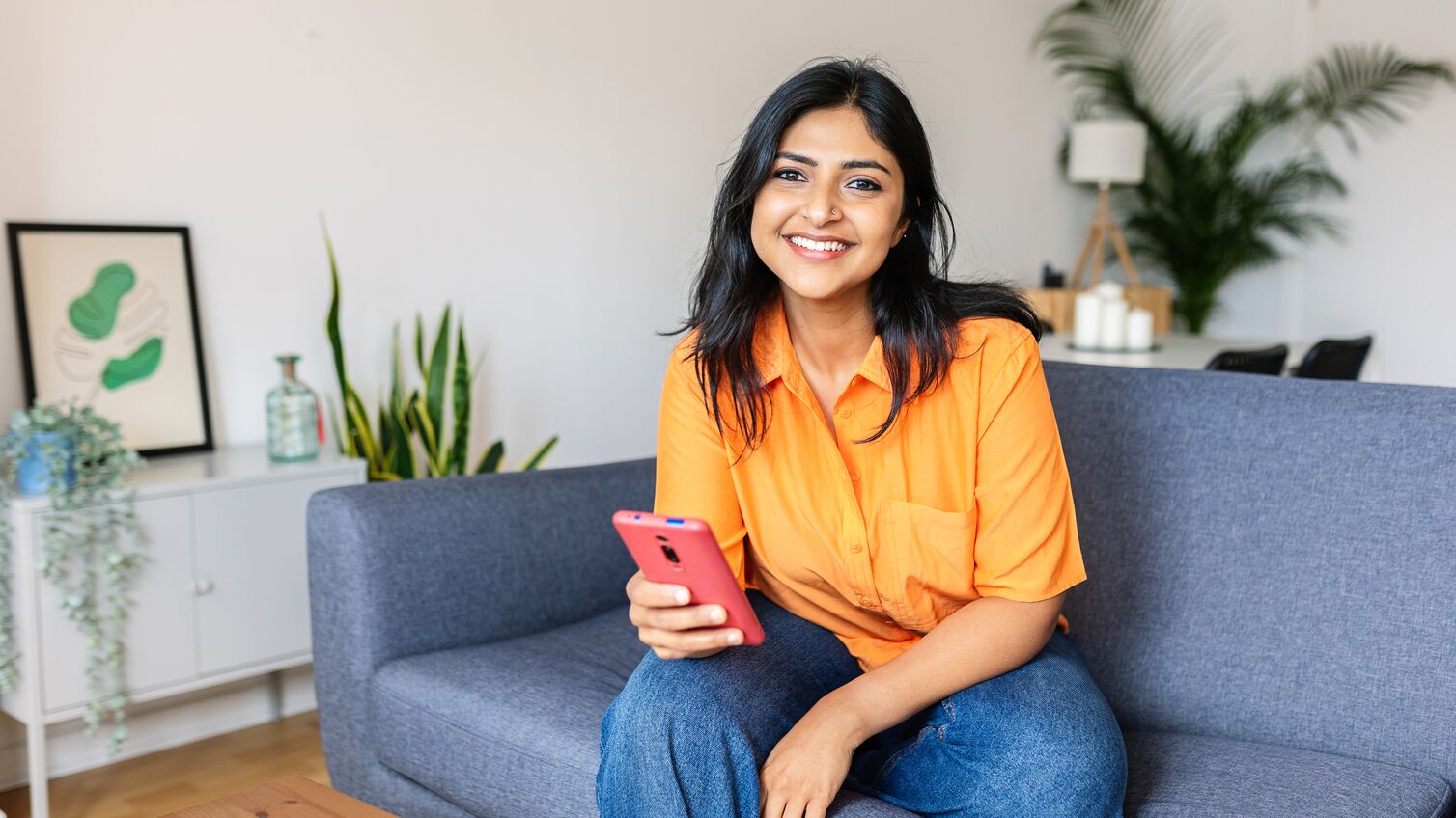 Candid portrait of happy young adult Indian woman holding smart phone device sitting at comfortable couch at home.