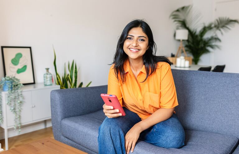 Candid portrait of happy young adult Indian woman holding smart phone device sitting at comfortable couch at home.