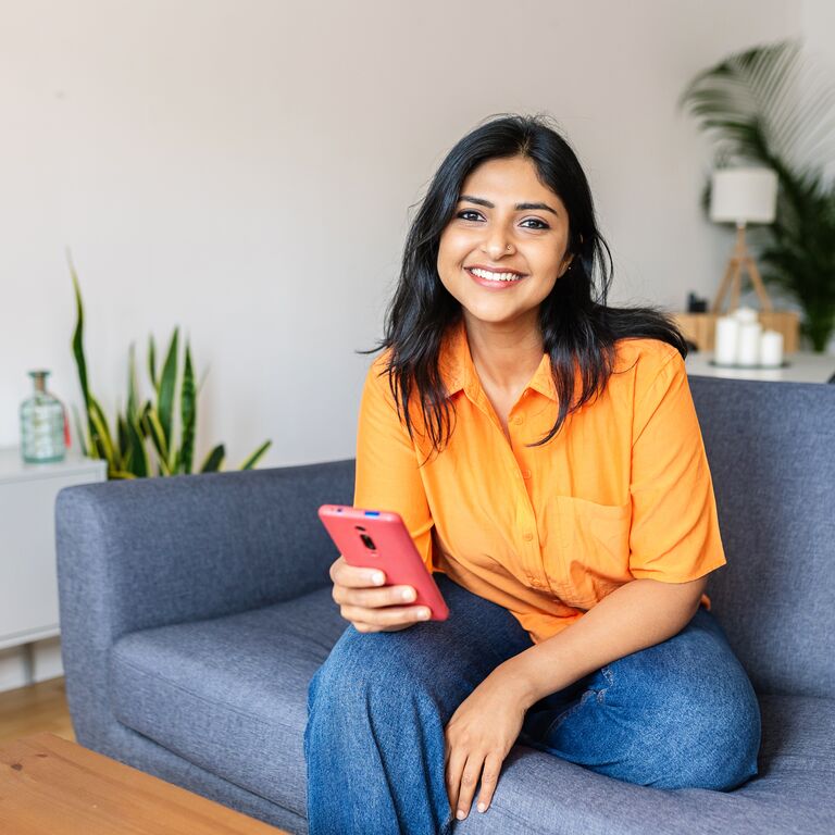 Candid portrait of happy young adult Indian woman holding smart phone device sitting at comfortable couch at home.