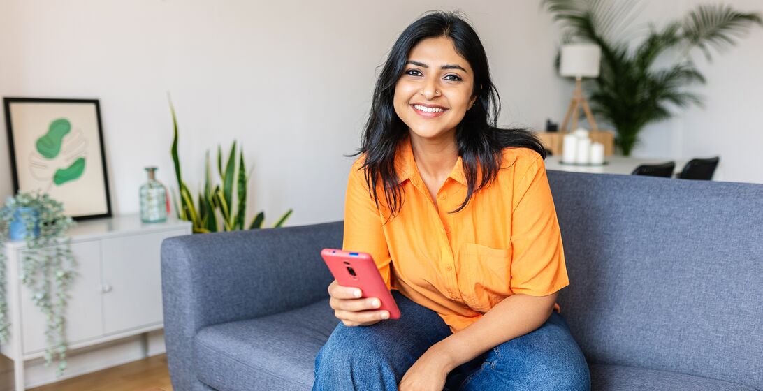 Candid portrait of happy young adult Indian woman holding smart phone device sitting at comfortable couch at home.