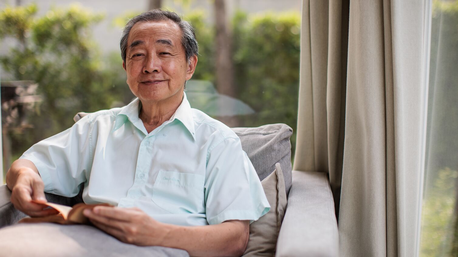 Portrait of a senior Thai man is seen reading a book in a bright and airy room. Seated in a comfortable armchair, the natural light streaming through the large glass windows enhances the calm and relaxing setting.