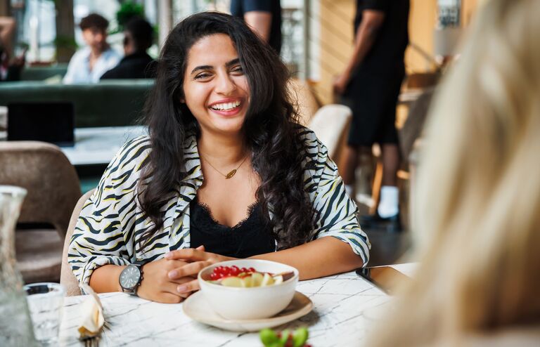 A cheerful woman with a radiant smile enjoys a bowl of fresh fruit in a vibrant café setting, exemplifying a moment of urban leisure and healthy living.