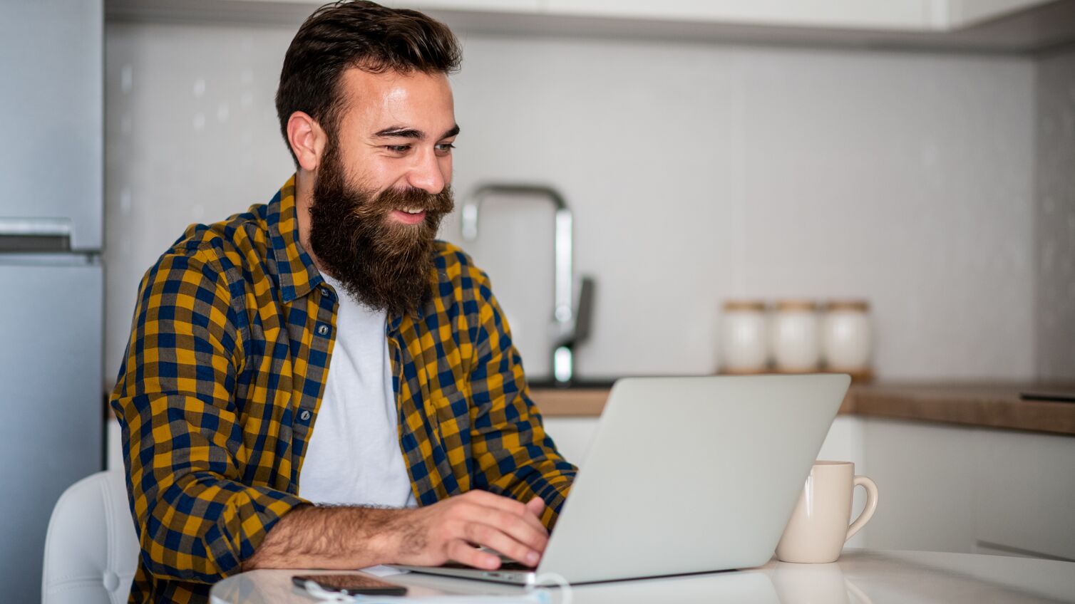 One young man is sitting in kitchen and typing on his laptop. He is freelancer and he works from home.