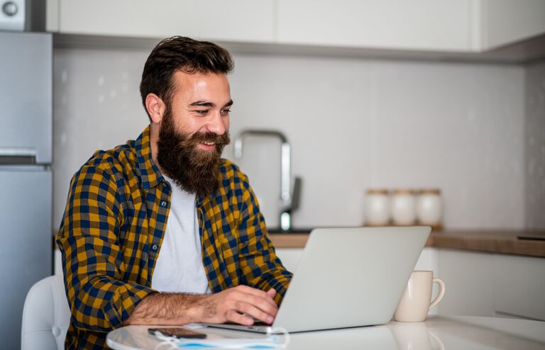 One young man is sitting in kitchen and typing on his laptop. He is freelancer and he works from home.