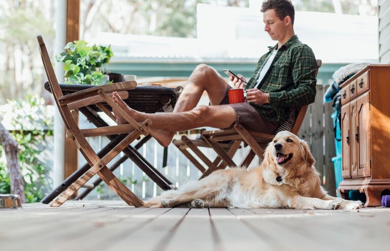 A side view shot of a young man sitting down on a chair outside his house using his smart phone with his dog.