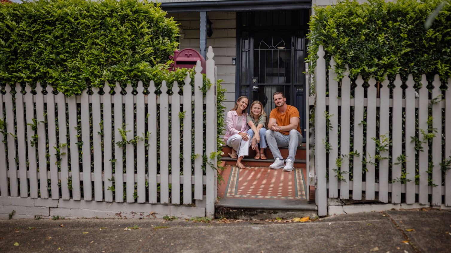 The family poses affectionately on the front steps of their house, surrounded by greenery and vibrant details, illustrating unity, love, and cheerful living.