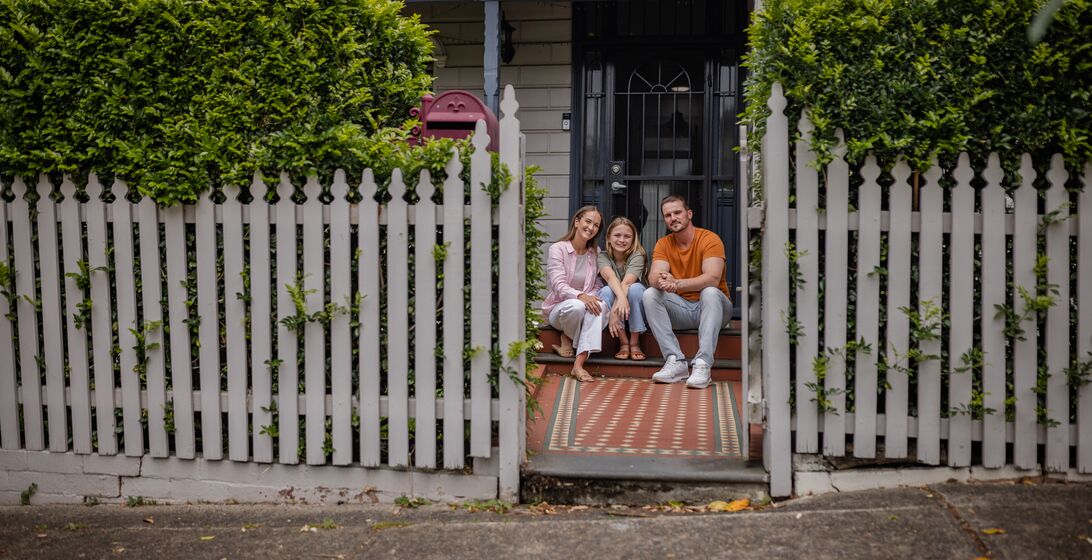 The family poses affectionately on the front steps of their house, surrounded by greenery and vibrant details, illustrating unity, love, and cheerful living.
