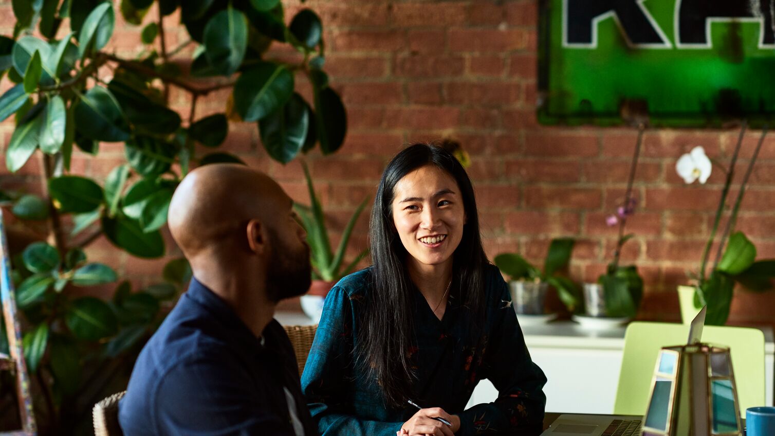 Two creative designers sitting in meeting in modern office, woman with note paper and pen listening to man with cheerful expression