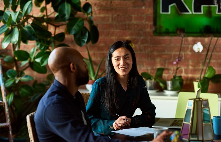 Two creative designers sitting in meeting in modern office, woman with note paper and pen listening to man with cheerful expression