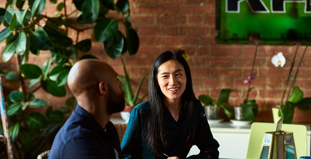 Two creative designers sitting in meeting in modern office, woman with note paper and pen listening to man with cheerful expression