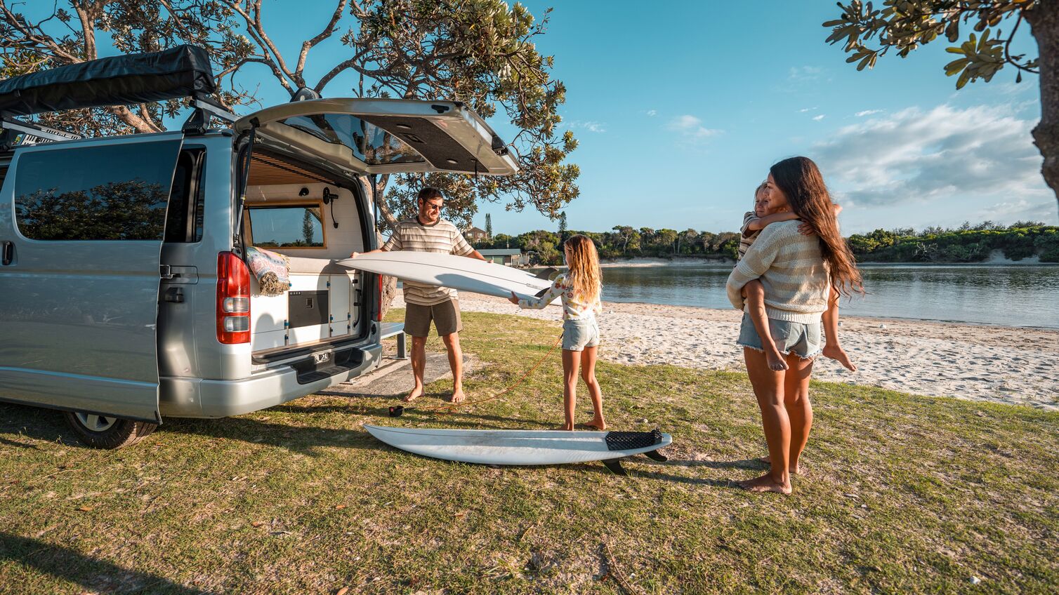 A Caucasian father unloads surfboards from a van during a family camping trip near a sunny beach, with his wife and daughters enjoying the serene outdoor setting.