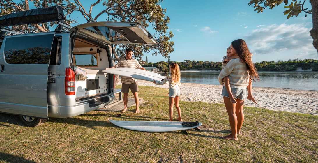 A Caucasian father unloads surfboards from a van during a family camping trip near a sunny beach, with his wife and daughters enjoying the serene outdoor setting.