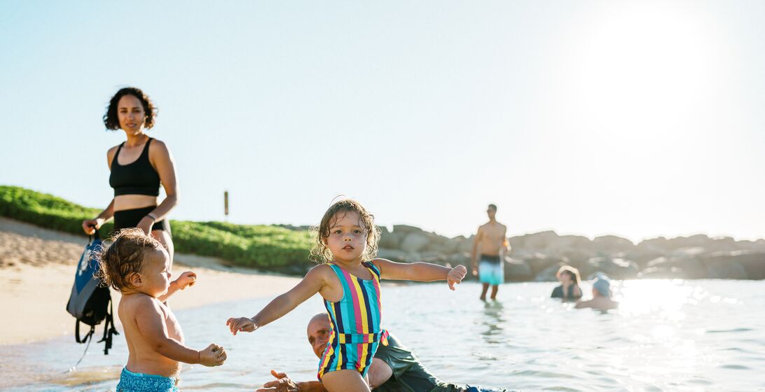 Two multiracial toddler children stand in the shallow ocean, swimming with their mother and grandfather during a tropical family vacation.