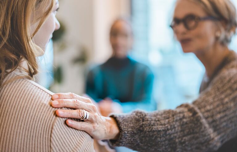 Senior woman comforting a female with hand on her shoulder during group therapy session. Close-up of a woman getting psychological support during therapy session