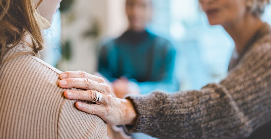 Senior woman comforting a female with hand on her shoulder during group therapy session. Close-up of a woman getting psychological support during therapy session