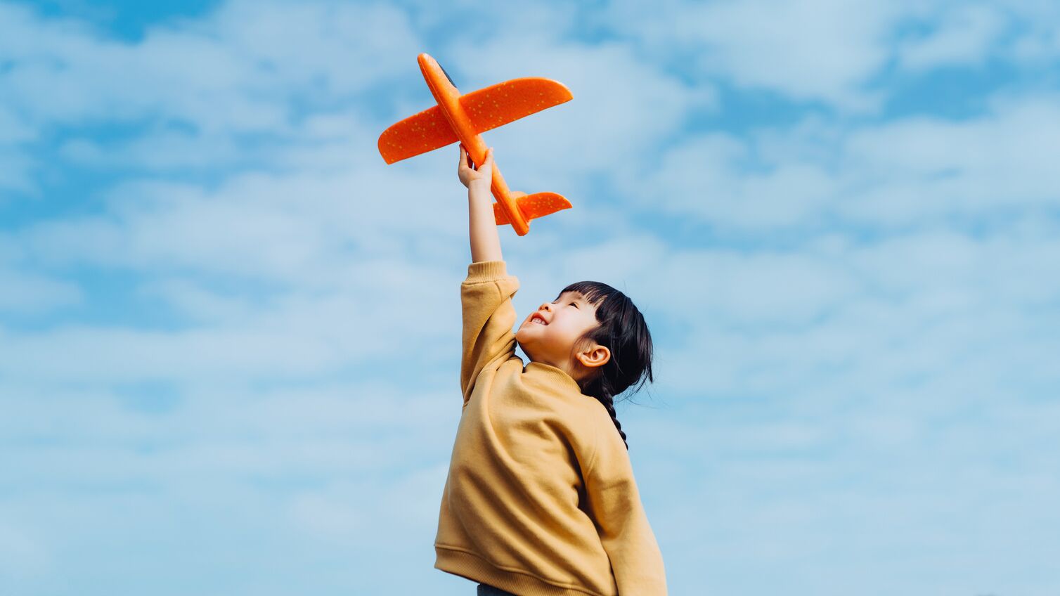 Happy lovely little Asian girl having fun outdoors, playing with airplane toy and smiling joyfully in park on a lovely sunny day against beautiful blue sky