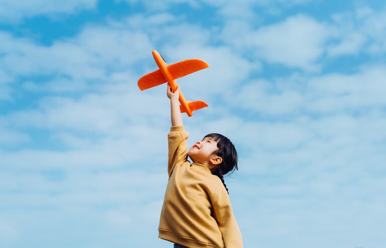 Happy lovely little Asian girl having fun outdoors, playing with airplane toy and smiling joyfully in park on a lovely sunny day against beautiful blue sky