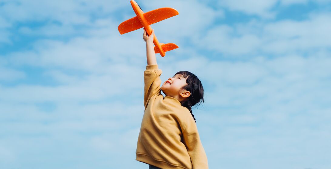 Happy lovely little Asian girl having fun outdoors, playing with airplane toy and smiling joyfully in park on a lovely sunny day against beautiful blue sky