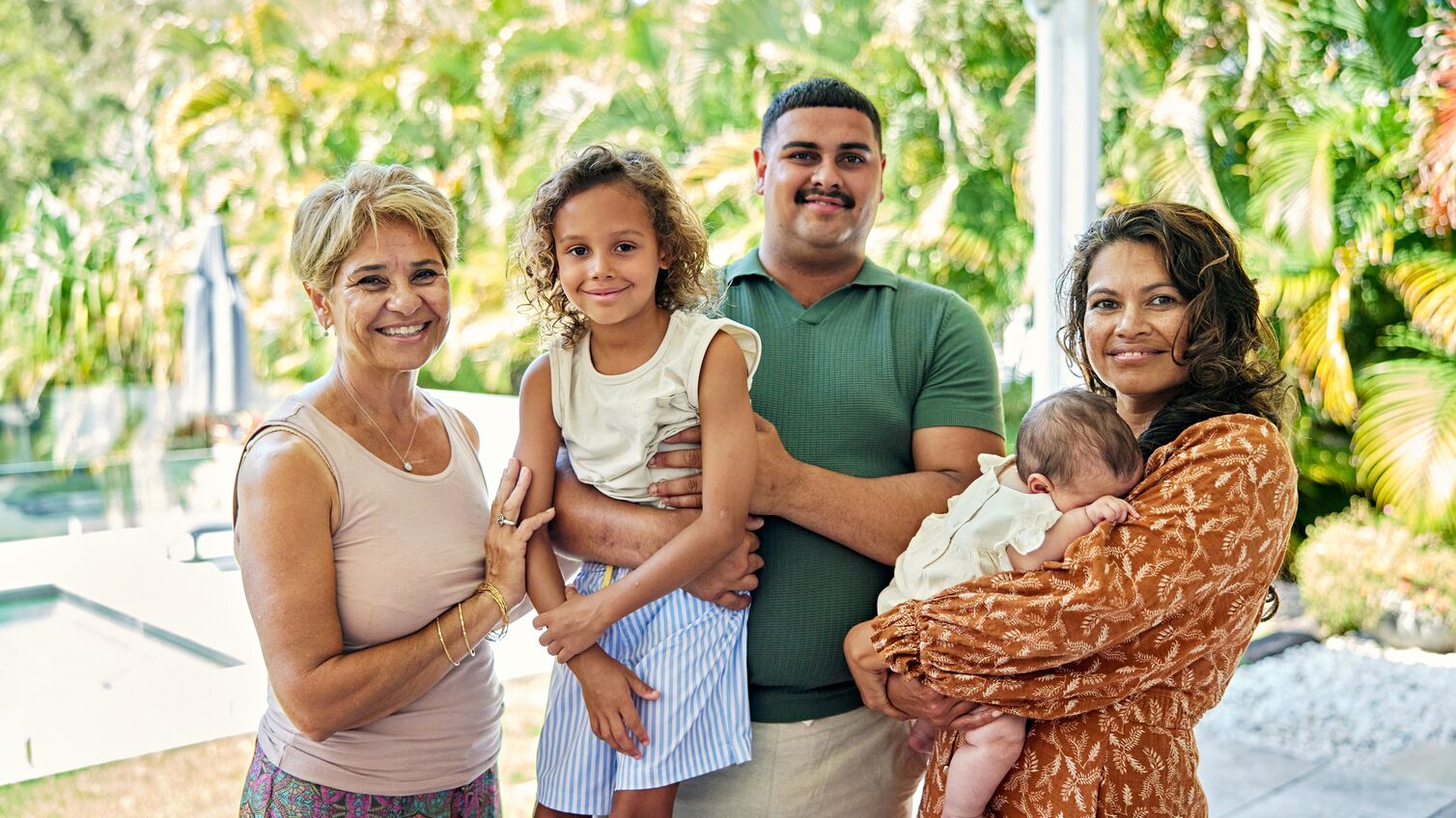 Casually dressed women in 40s and 60s, man in 20s holding 7 year old boy and 3 month old baby in arms, everyone smiling at camera, sunny foliage in background.