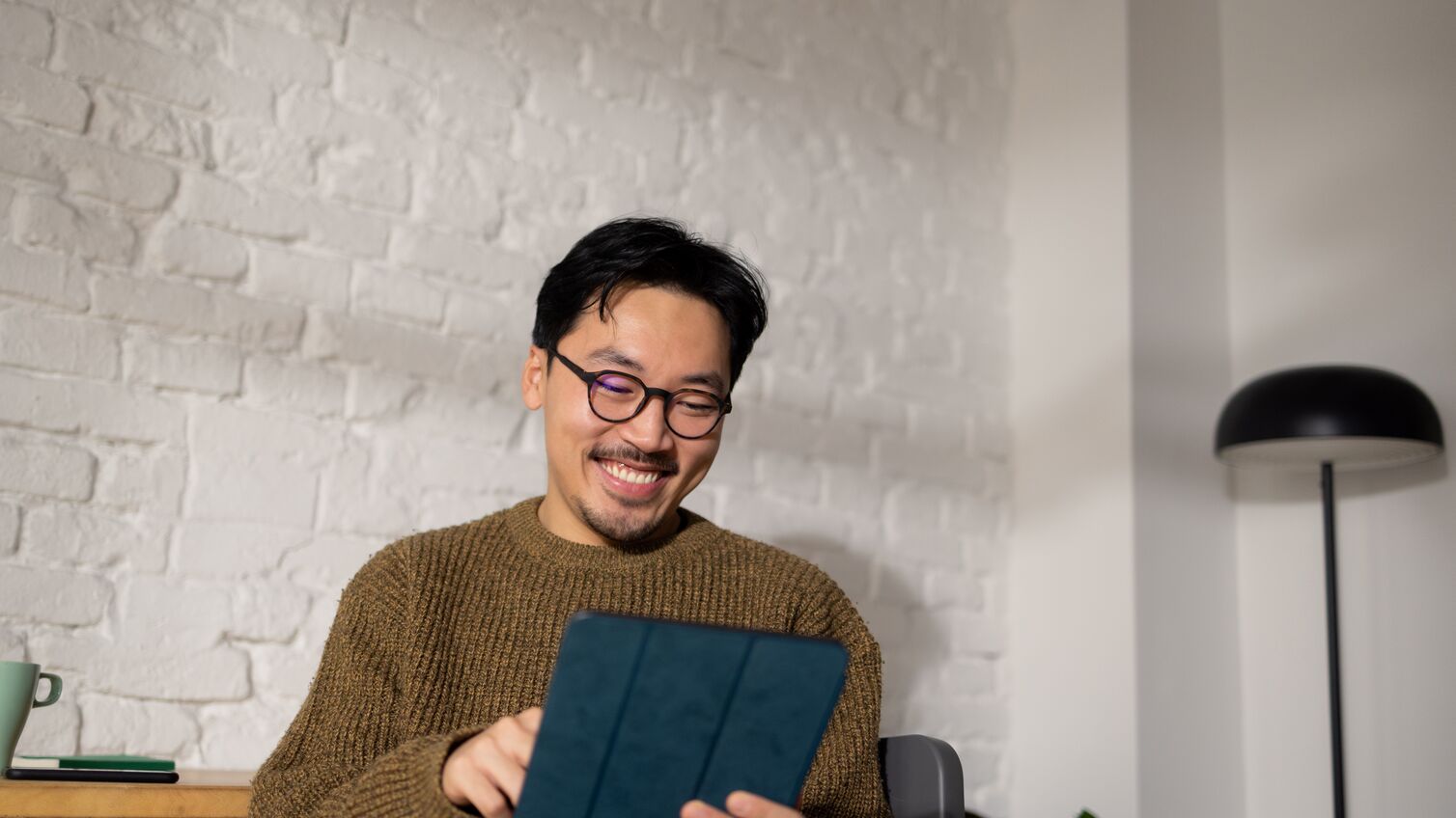 Young hip Chinese man sitting at his home office while enjoying his work using tablet