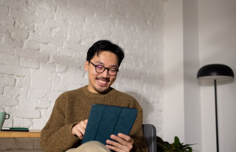 Young hip Chinese man sitting at his home office while enjoying his work using tablet