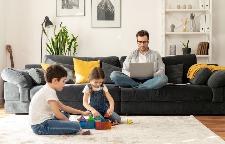 Father using laptop for freelance work sitting on the sofa at home while his two kids playing peacefully together on the floor. Busy single parent is babysitting and working on the distance
