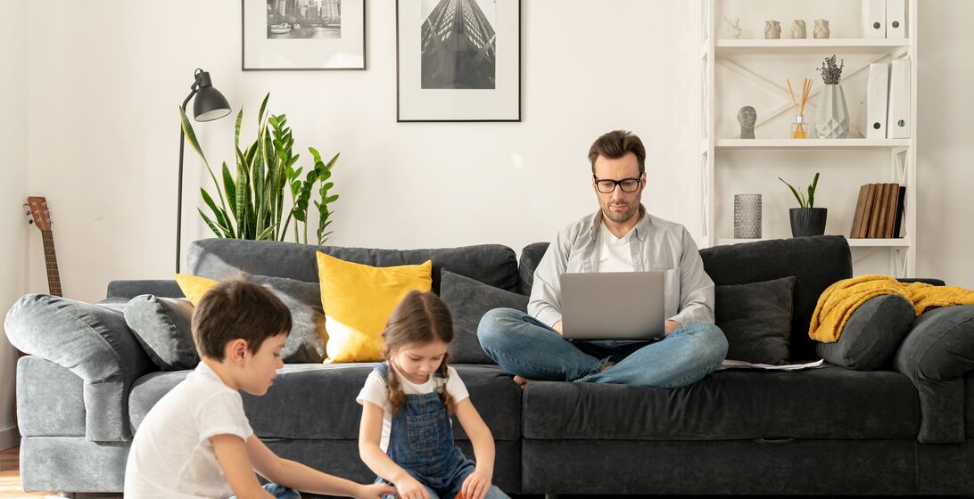 Father using laptop for freelance work sitting on the sofa at home while his two kids playing peacefully together on the floor. Busy single parent is babysitting and working on the distance