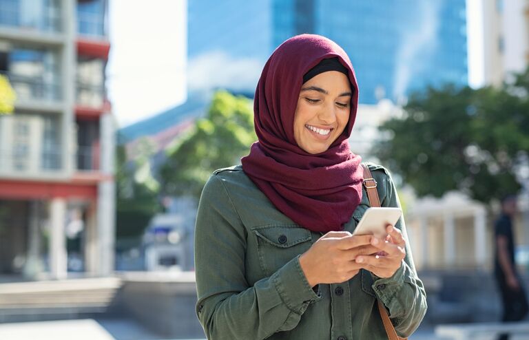 Pretty muslim woman using mobile phone outdoor. Arabic woman wearing hijab using smartphone on the street. Islamic girl texting a message phone in city centre.