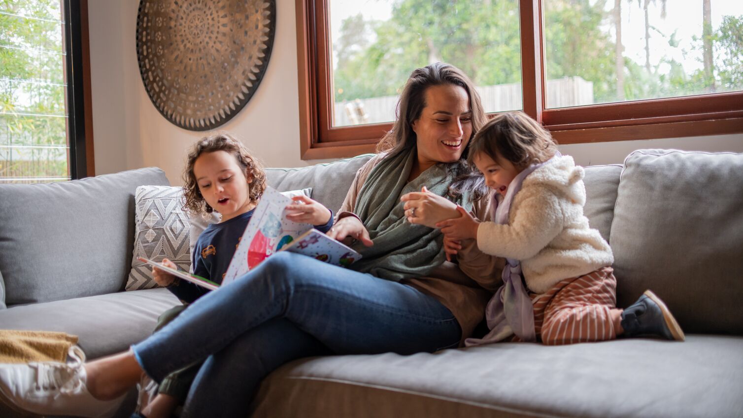 A mother laughs and plays with her two young children. They are snuggled up on the couch together and enjoying quality family time.