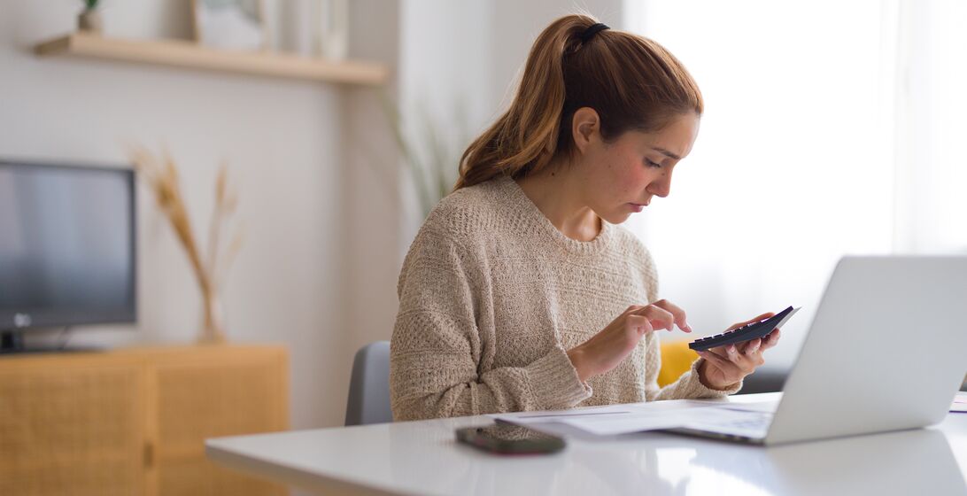 Focused young woman managing home finances, calculating bills and expenses using calculator and laptop in her living room