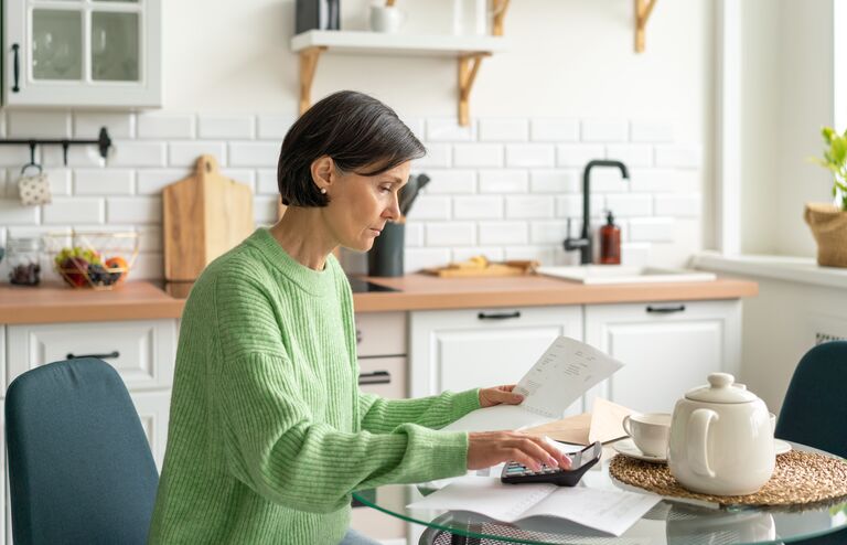 Senior adult woman handling finance from home. Concept of paying bills and doing taxes in kitchen. Wisely planning personal budget and calculating savings. Focused on paperwork while working at home. Engaging with home finances remotely