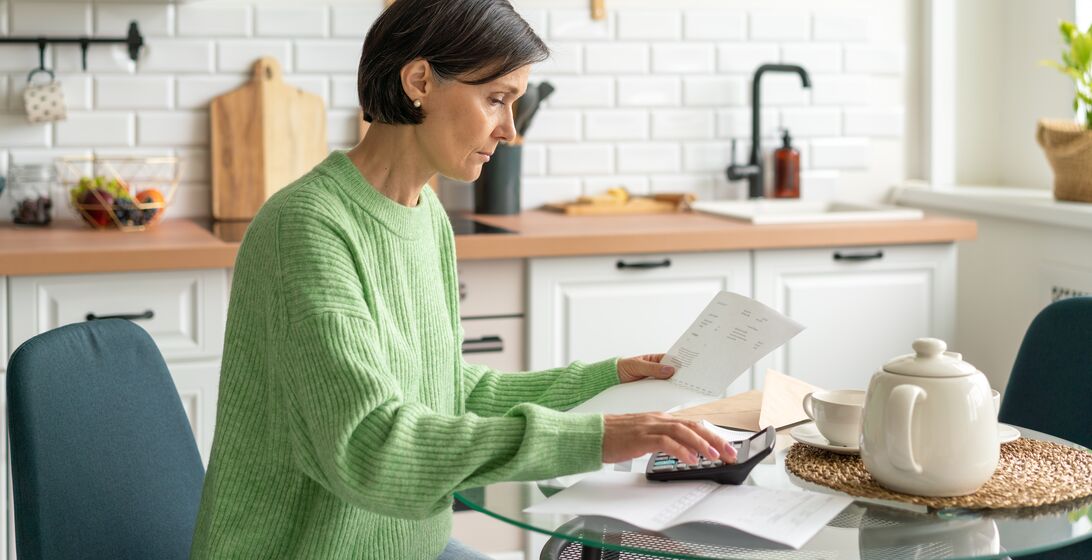 Senior adult woman handling finance from home. Concept of paying bills and doing taxes in kitchen. Wisely planning personal budget and calculating savings. Focused on paperwork while working at home. Engaging with home finances remotely