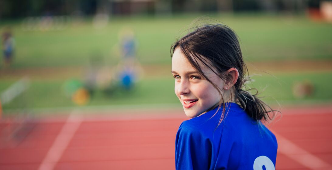 Little girl is watching over her shoulder while sitting by the running track at athletics club.