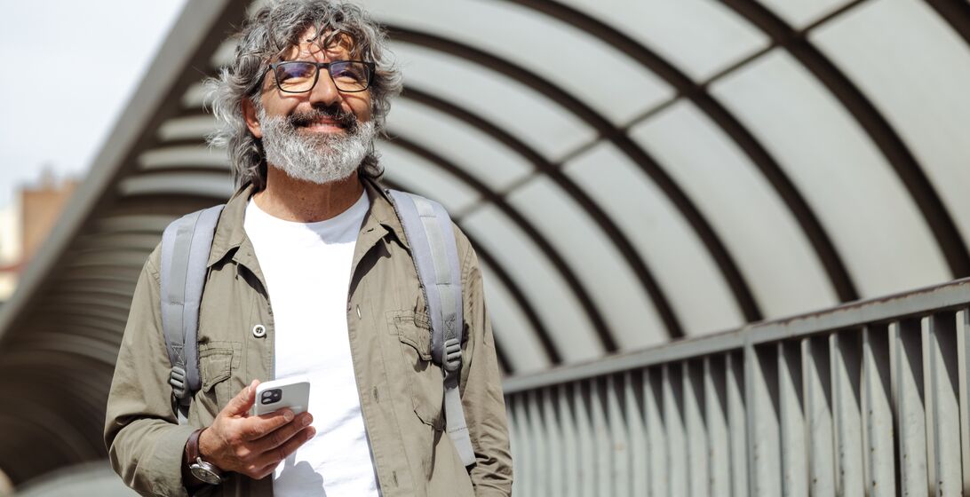 A modern senior man is walking on the street and smiling. He is holding a smart phone and smiling