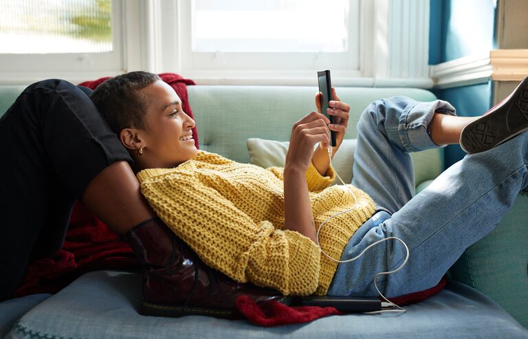 Smiling woman text messaging on smart phone while leaning on friend's leg in living room