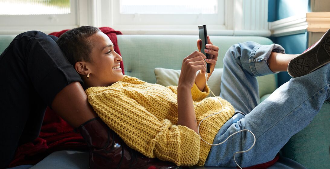 Smiling woman text messaging on smart phone while leaning on friend's leg in living room