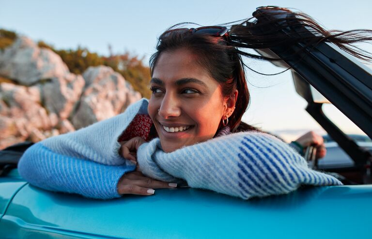 Smiling young woman day dreaming while leaning on convertible car during road trip