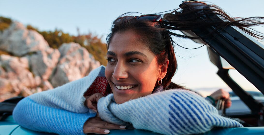 Smiling young woman day dreaming while leaning on convertible car during road trip