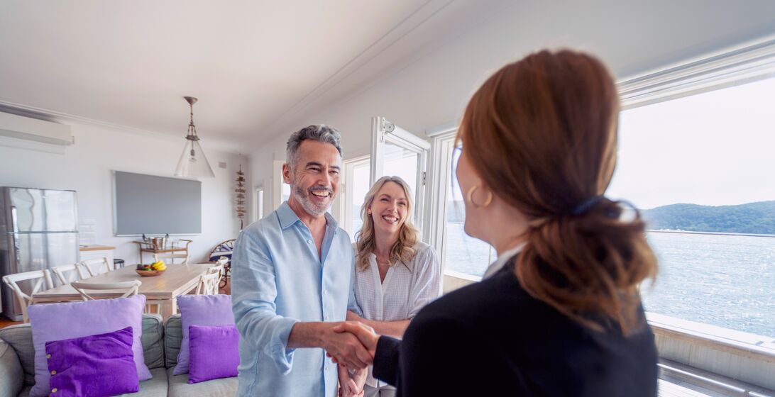 Real estate agent showing a mature couple a new house. The house is contemporary. All are happy and smiling. The couple are casually dressed and the agent is in a suit. The property is waterfront with the sea in the background.