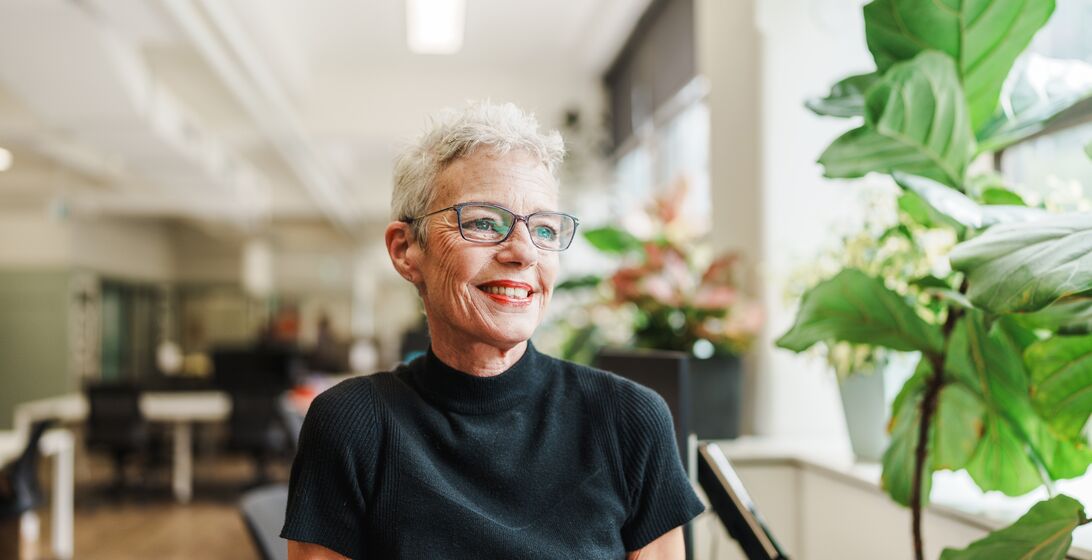 Senior woman wearing a black short sleeved turtleneck and glasses, sitting in an office.