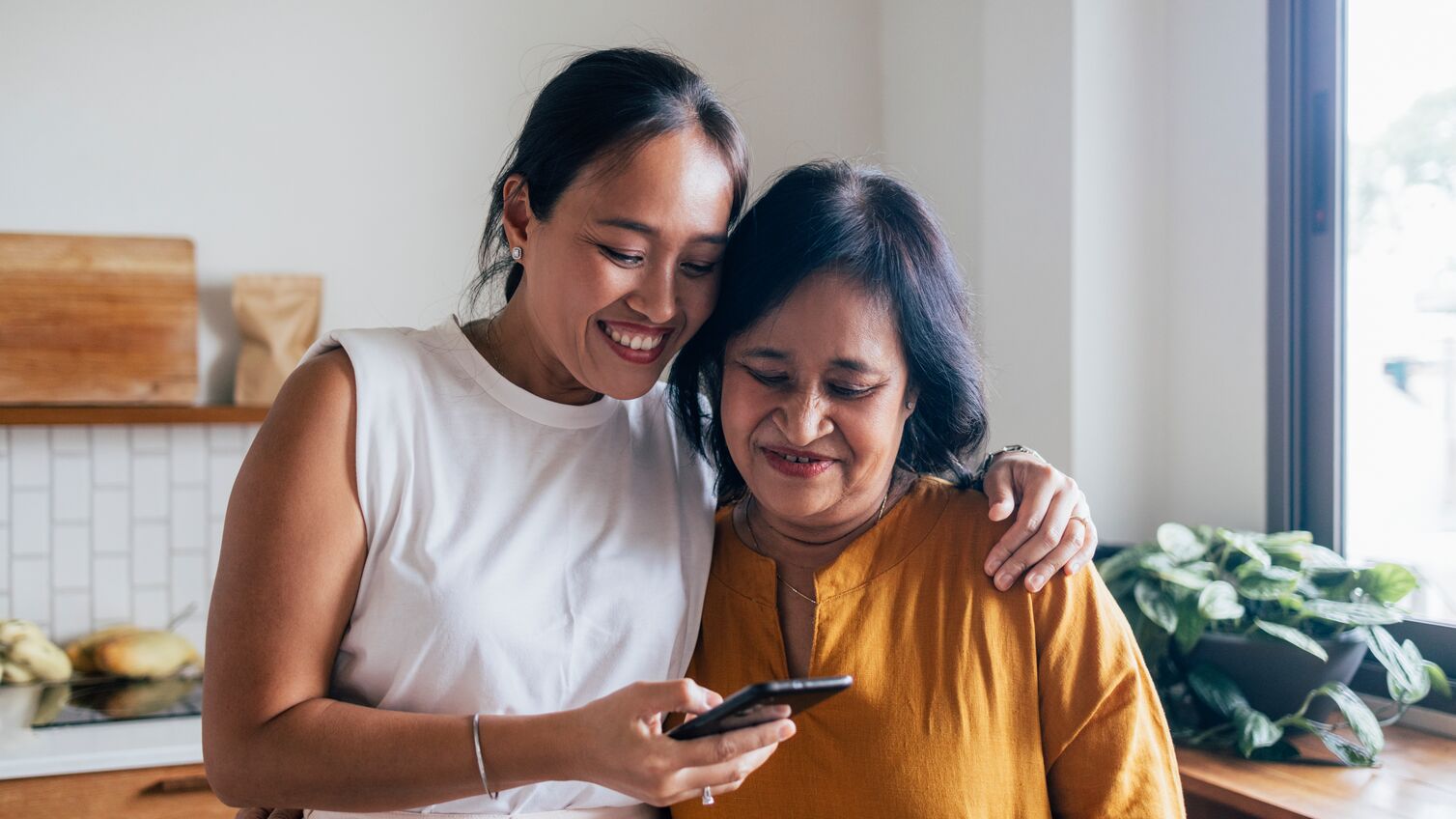 A smiling Asian female hugging her mother while they are reading something online using their black smartphone.